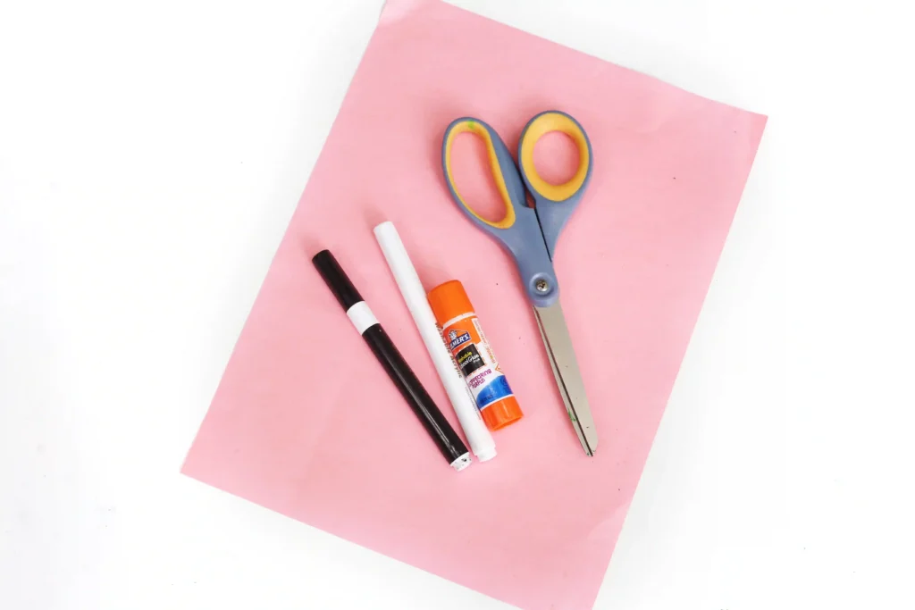 Craft supplies for the pink bat handprint craft on pink paper: blue-handled scissors, markers and glue stick ready for a DIY project outside the frame edge.