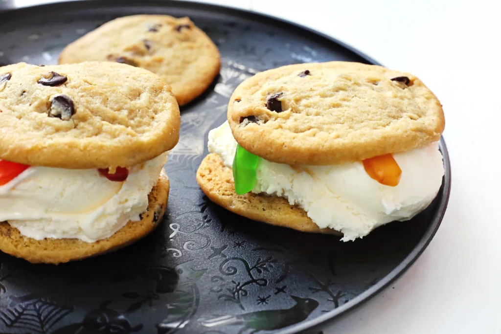 Close-up of two chocolate chip cookies with a vanilla ice cream in the middle with gummy worms on a dark decorative plate