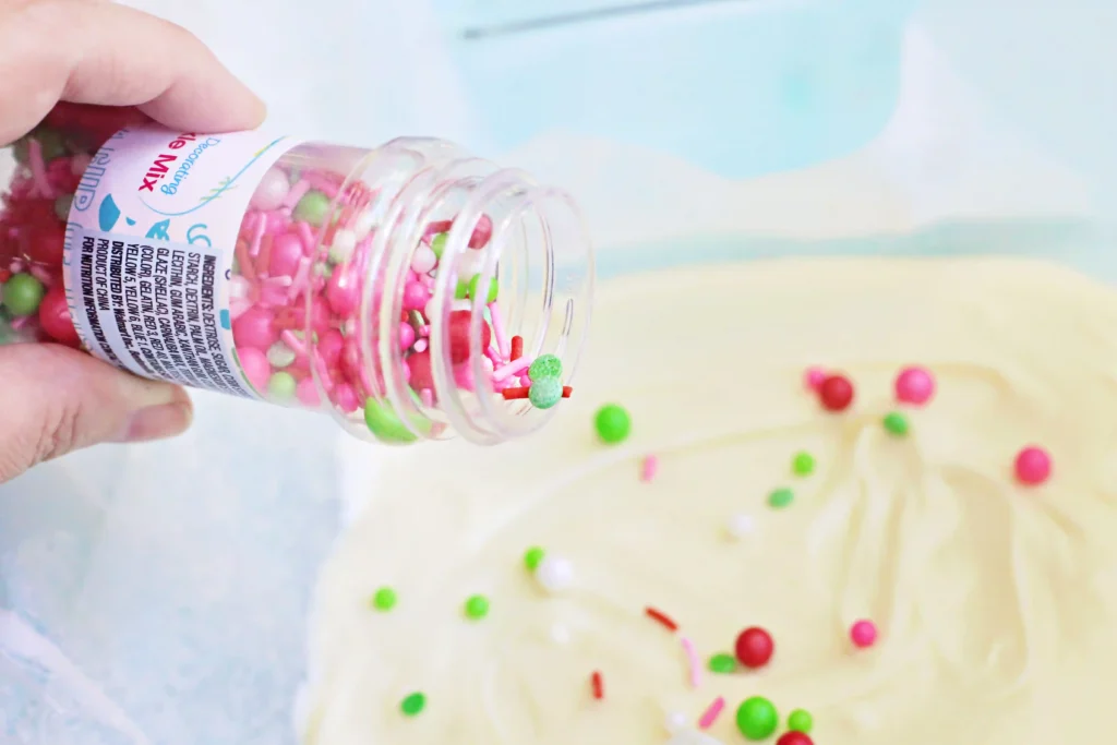 Person tilting a jar to pour pink and green candy sprinkles onto melted white chocolate.