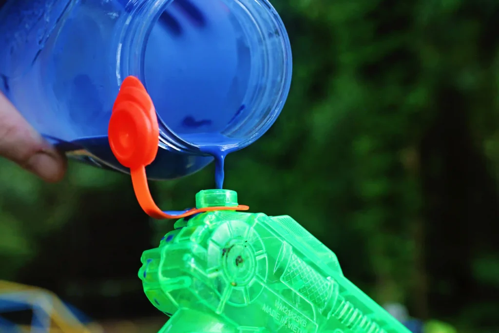 Close-up of a blue bottle pouring bright blue paint into a green toy water gun, with an orange nozzle in the foreground outdoors.