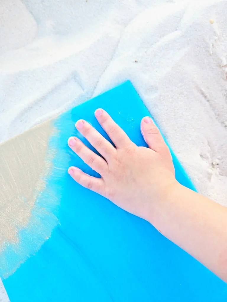 Child's hand presses a bright blue canvas board on light sand.