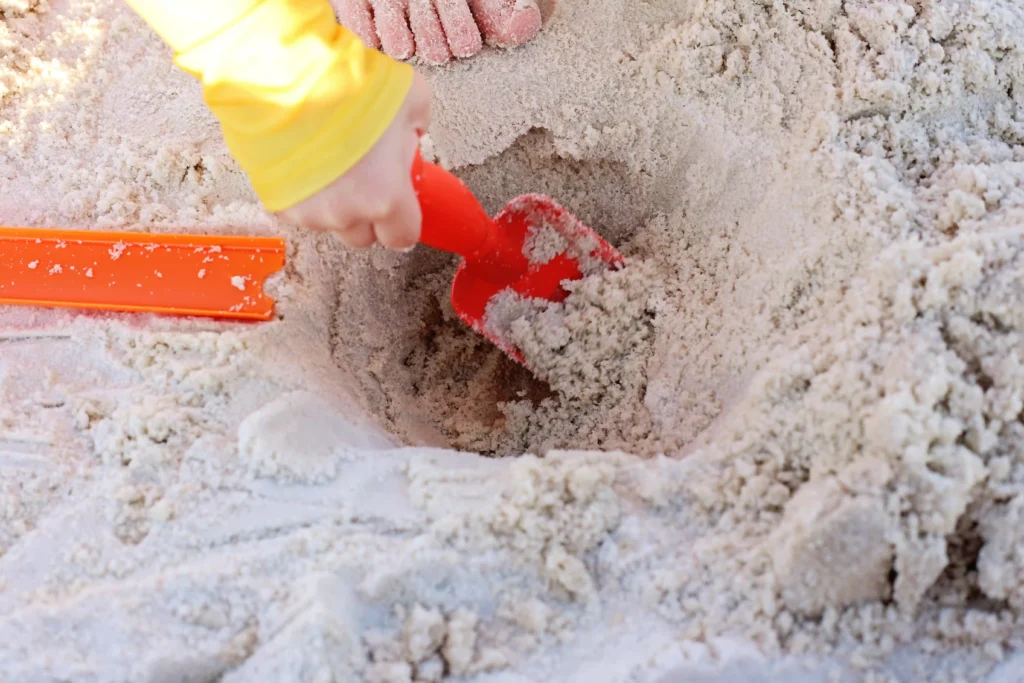 Child uses a red plastic shovel to dig  a hole in the sand