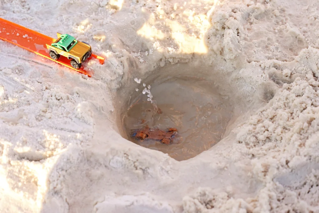 Toy jeep on an orange shovel at the edge of a snowy hole, with small orange figures visible in the water below.