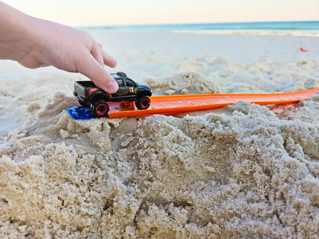 Small toy pickup truck on an orange beach ramp in wet sand, with the ocean in the distance.