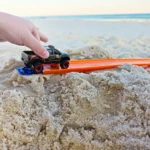 Small toy pickup truck on an orange beach ramp in wet sand, with the ocean in the distance.