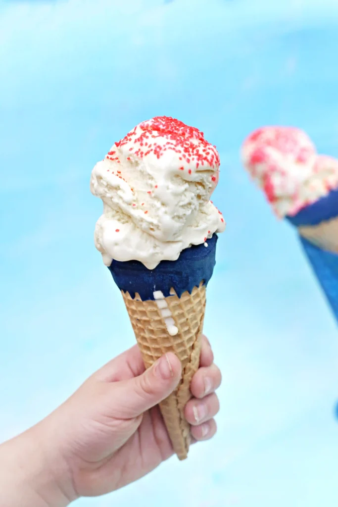A hand holding a blue dipped waffle cone with vanilla ice cream topped with red sprinkles against a light blue background to make a patriotic ice cream cone.