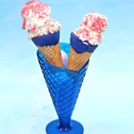 Two ice cream cones dipped in blue chocolate and white ice cream topped with red sprinkles in a blue holder against a pale blue background