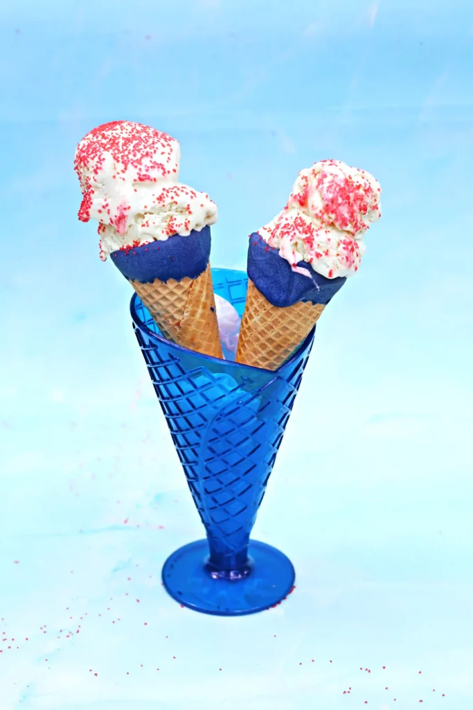 Two ice cream cones dipped in blue chocolate and white ice cream topped with red sprinkles in a blue holder against a pale blue background