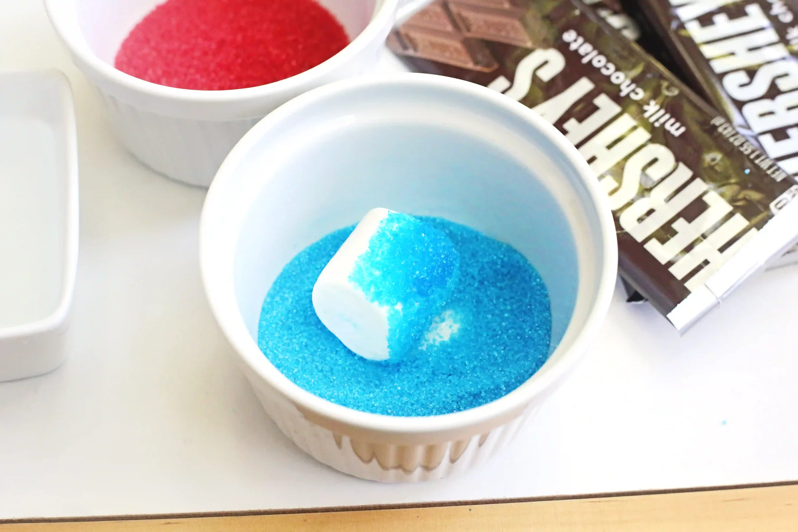 Two white cups hold bright colored sugar—blue in the front, red in the back—with a white spoon scooping blue sugar.