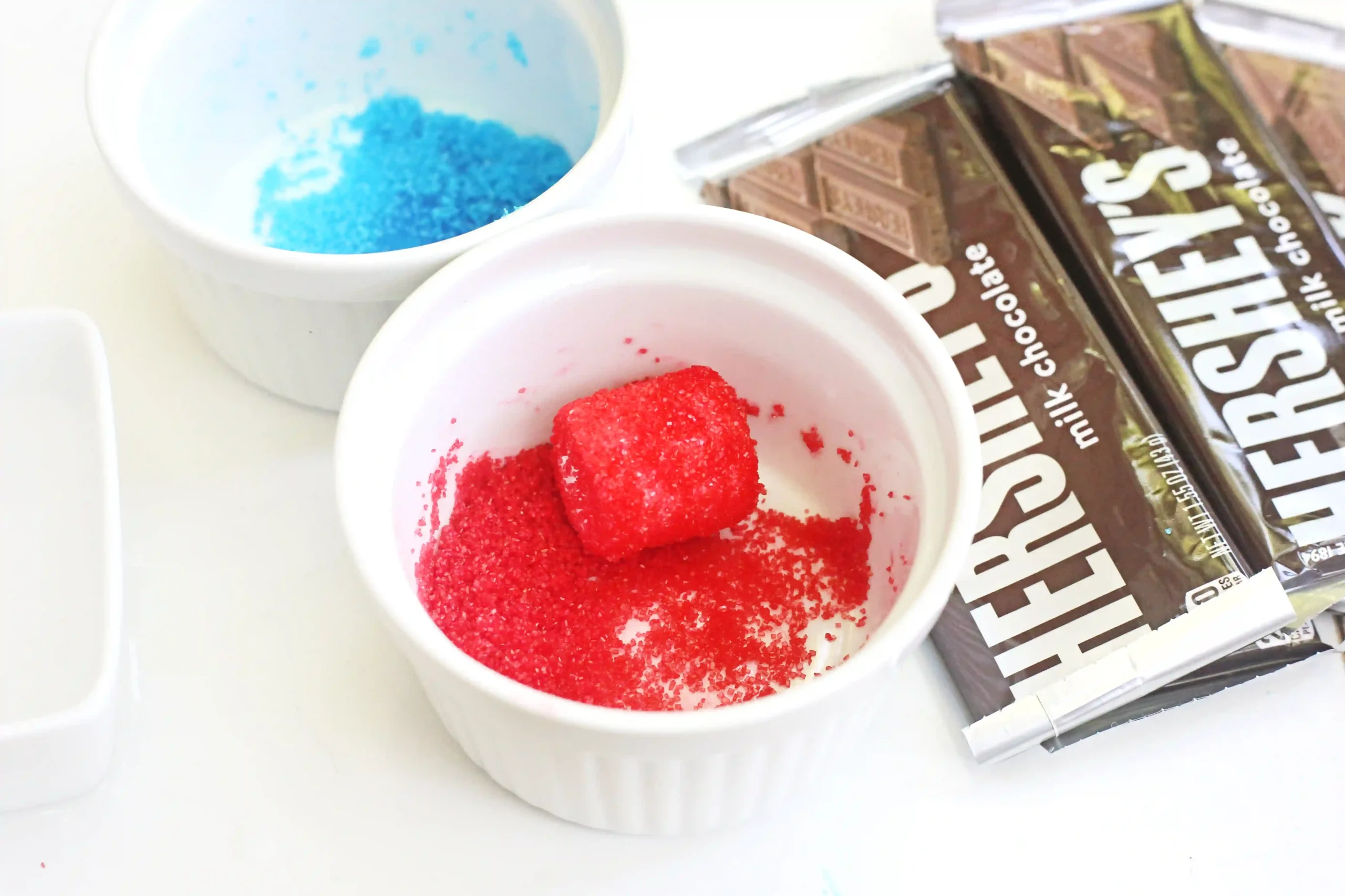 White cups contain blue and red sugar crystals placed beside a chocolate bar wrapper; a colorful confection setup ready for decorating desserts.
