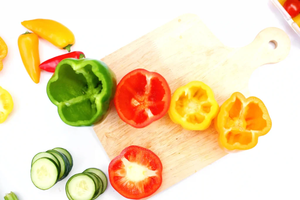 Assorted bell peppers (green, red, yellow) sliced on a wooden cutting board with cucumber rounds nearby on a white surface.