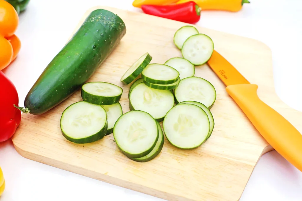 Cucumber slices on a wooden cutting board with an orange knife nearby; colorful peppers visible in the background.