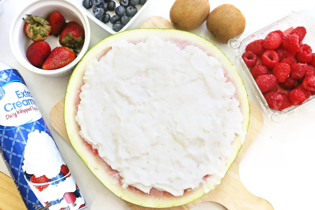 Watermelon half topped with whipped cream surrounded by berries (strawberries, raspberries, blueberries) and kiwi/kiwi pieces nearby; whipped topping tube in foreground.