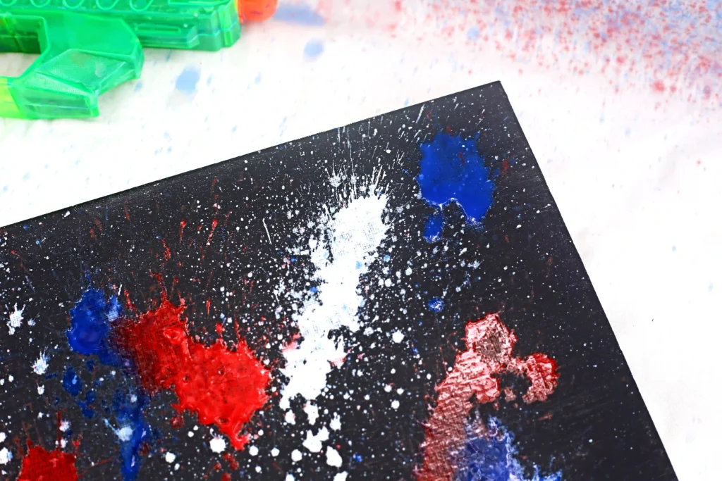 Close-up of an abstract paint-splattered black canvas with red, blue, and white splashes; green translucent art supplies in the top-left corner