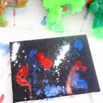 Neon-green and orange toy water guns on a paint-splattered surface beside a black canvas with red, blue, and white splatters.
