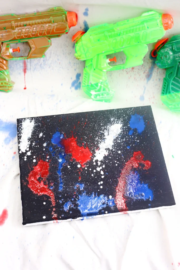 a squirt gun firework painting : Neon-green and orange toy water guns on a paint-splattered surface beside a black canvas with red, blue, and white splatters.
