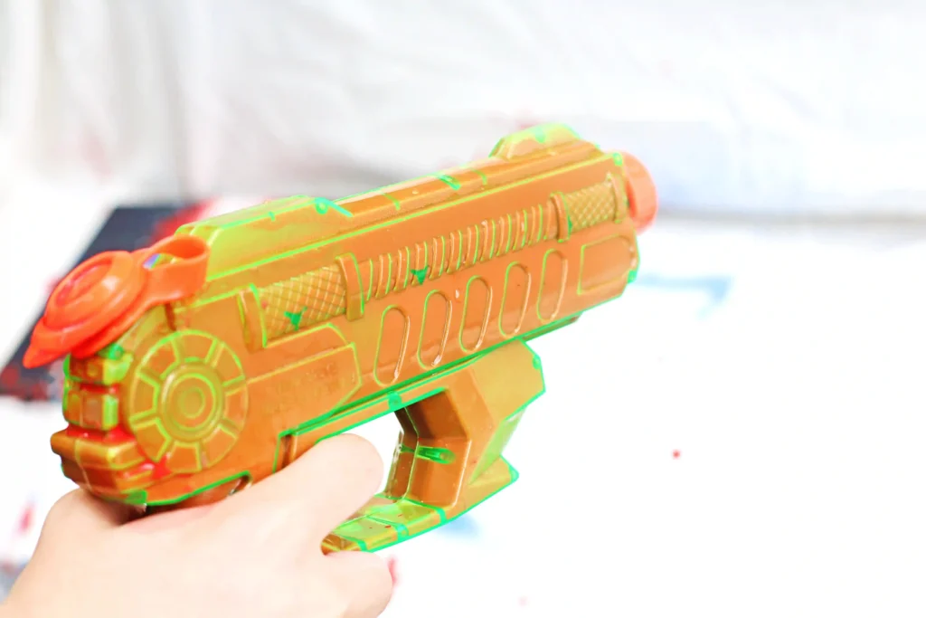 Close-up of a bright orange squirt gun filled with paint with green accents being held by a hand.