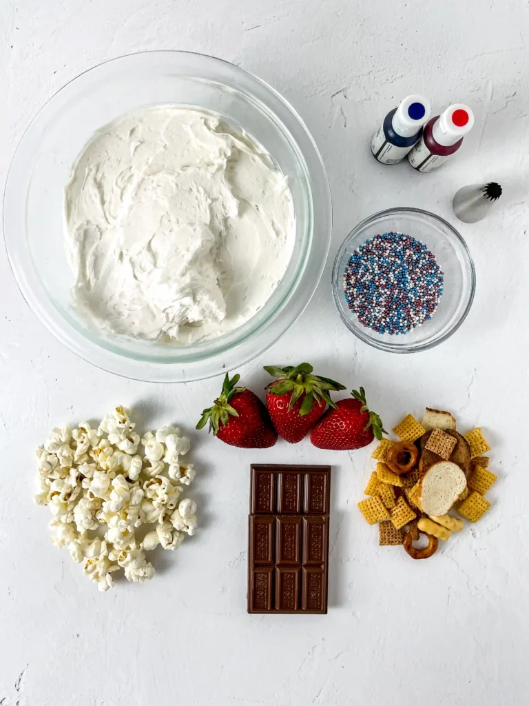 ingredients for the patriotic charcuterie board - Top-down view of a baking setup: bowl of white frosting, chocolate bar, strawberries, popcorn, sprinkles, and coloring bottles on a white surface.