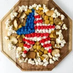 a patriotic dessert -Top-down view of a wooden hex board covered with red, white, and blue frosting rosettes, popcorn, and assorted crackers.