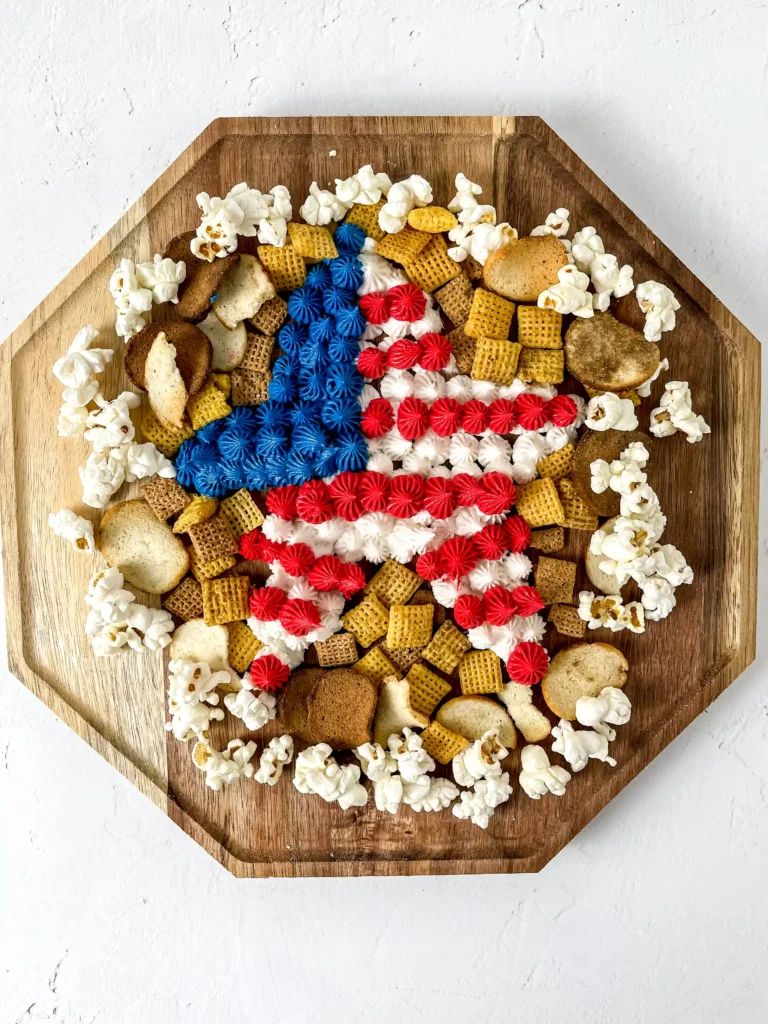 a patriotic dessert -Top-down view of a wooden hex board covered with red, white, and blue frosting rosettes, popcorn, and assorted crackers.