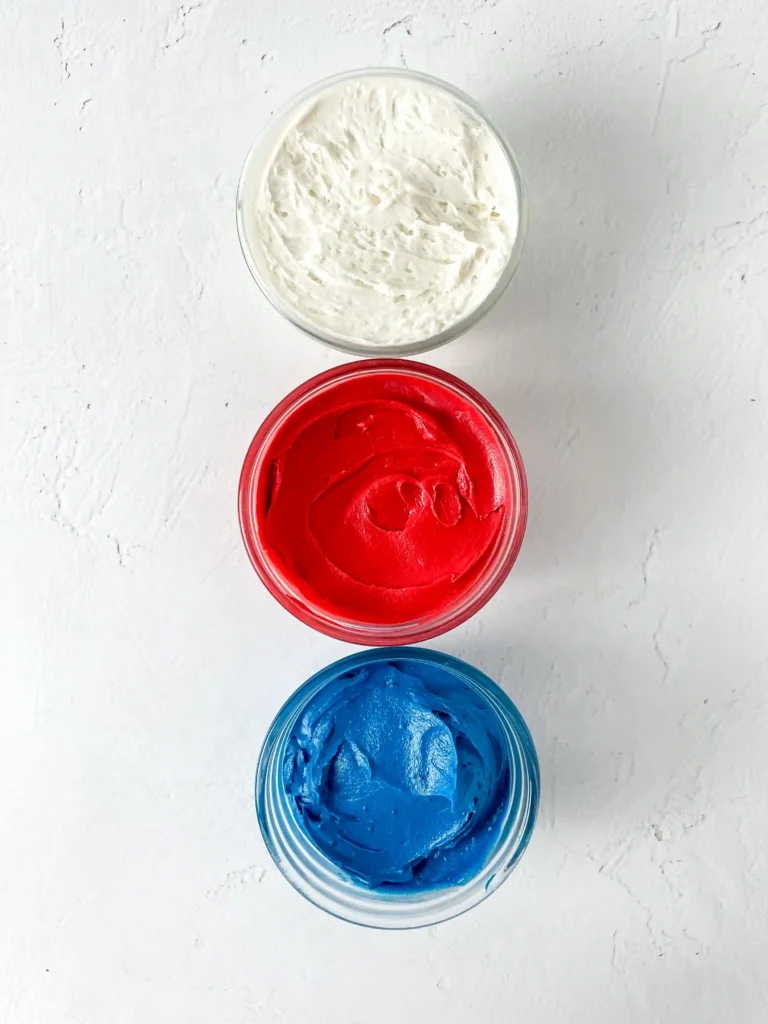 Three bowls of thick white, red, and blue buttercream frosting arranged vertically on a white textured surface