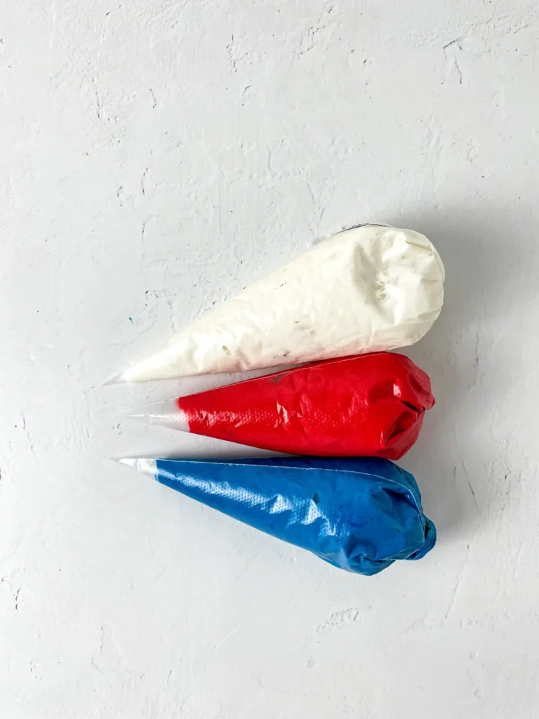 Three piping bags filled with icing in white, red, and blue resting on a textured white surface.