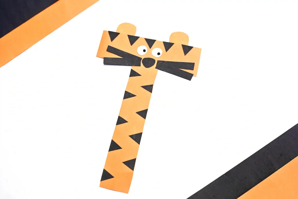 Paper tiger craft made from orange strips with black triangle patterns on a white surface, with diagonal caution stripes in the background