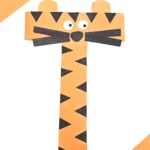 Orange paper tiger craft composed of strip-cut shapes with black triangular stripes, round white eyes, and a black nose to make up this letter activity