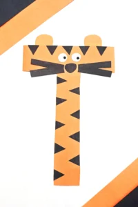 Orange paper tiger craft composed of strip-cut shapes with black triangular stripes, round white eyes, and a black nose to make up this letter activity