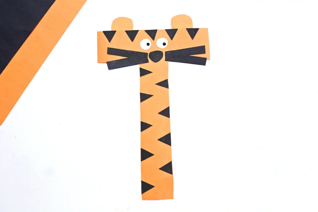 Orange paper tiger alphabet craft composed of strip-cut shapes with black triangular stripes, round white eyes, and a black nose.