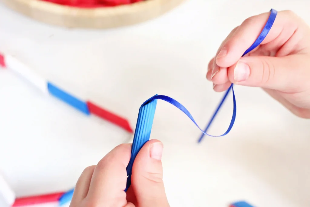 Patriotic Pasta Necklaces - Cultivating Brilliant Minds Close-up of hands threading blue pasta on a blue ribbon for this patriotic craft