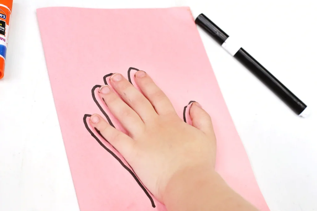 tracing of a hand with a black outline on pink paper with a black marker; glue stick visible on the left.