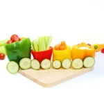 vegetables organized to look like a vegetable train that has various veggies inside bell peppers with cucumber slices for wheels