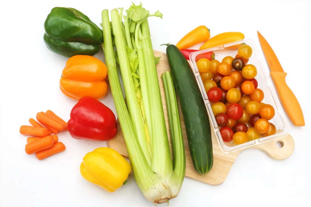 Assorted fresh vegetables on a cutting board: celery stalks, bell peppers (green, orange, yellow, red), a cucumber, carrot sticks, and cherry tomatoes in a clear container with a knife nearby.