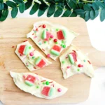 White chocolate bark topped with red-and-green watermelon gummies and colorful sprinkles on a wooden board, with green foliage above.
