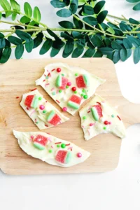 White chocolate bark topped with red-and-green watermelon gummies and colorful sprinkles on a wooden board, with green foliage above.