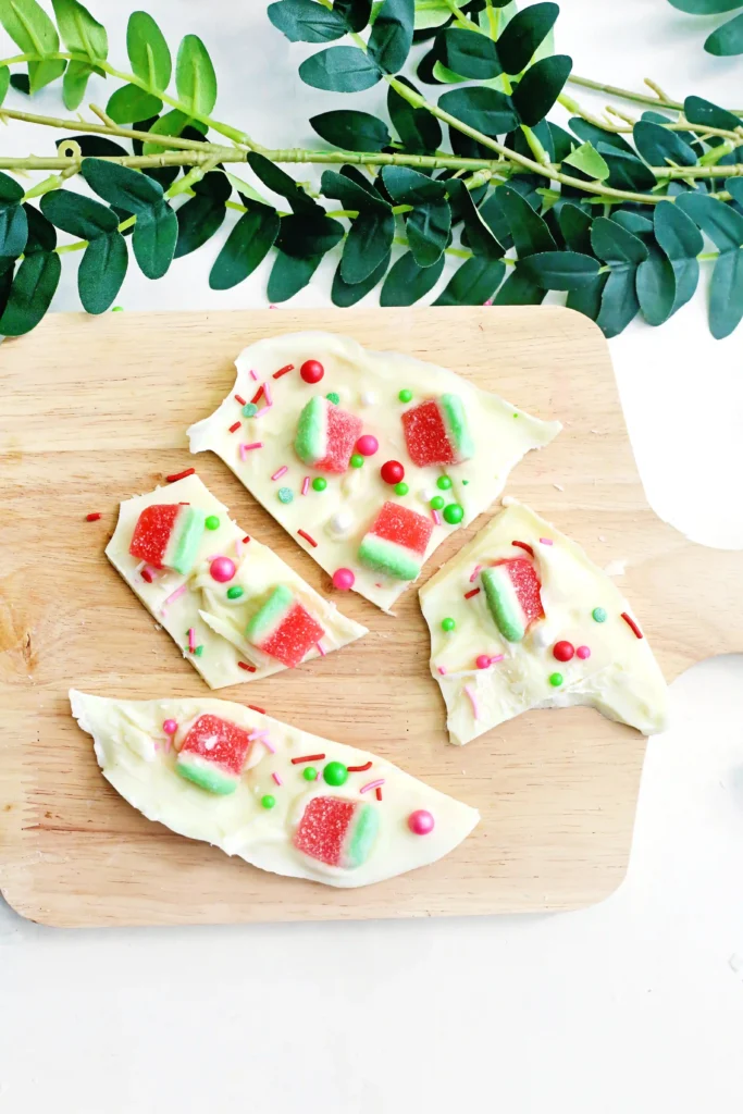 White chocolate bark topped with red-and-green watermelon gummies and colorful sprinkles on a wooden board, with green foliage above.