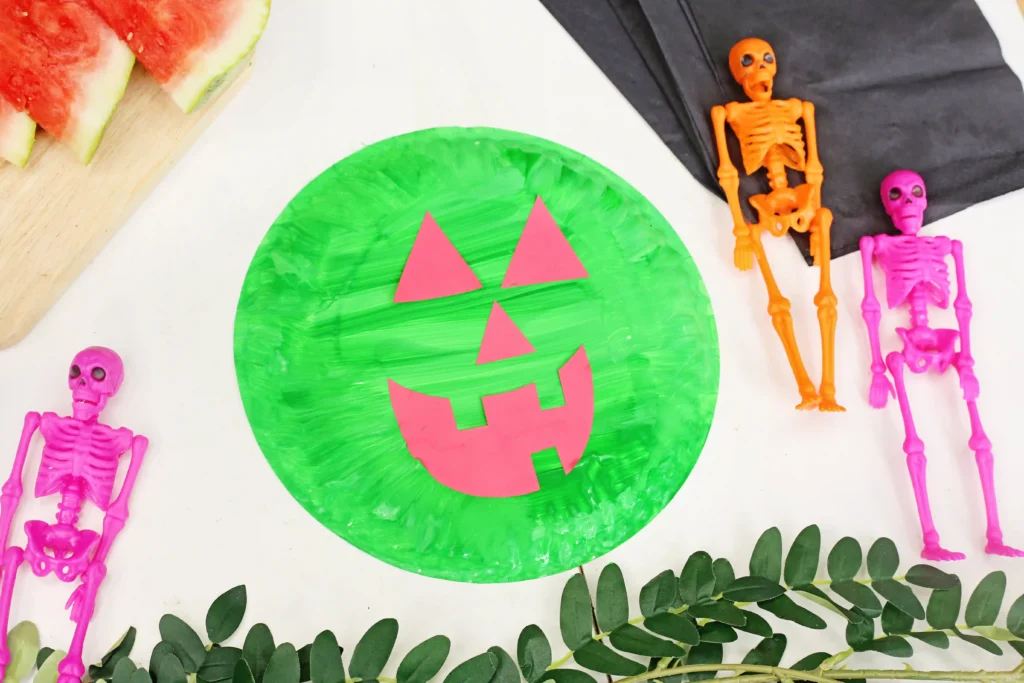 Bright green paper pumpkin with a pink jack-o'-lantern face sits on a white surface, surrounded by pink plastic skeletons, watermelon slices, and green leaves for a festive Halloween craft scene.