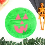 Green paper pumpkin with pink jack-o'-lantern face shapes, placed on a white surface with Halloween props nearby (orange skeleton, black napkins).