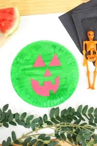 Green paper pumpkin with pink jack-o'-lantern face shapes, placed on a white surface with Halloween props nearby (orange skeleton, black napkins).