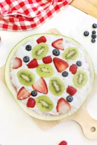 watermelon fruit pizza dessert on a melon rind crust topped with kiwi, strawberries, raspberries, and blueberries on a wooden board with a red checkered cloth in the background