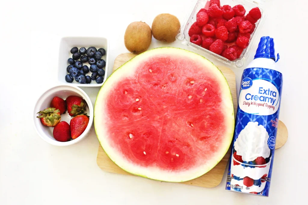 supplies for the watermelon pizza - Half watermelon on a cutting board surrounded by blueberries, strawberries, kiwis, raspberries, and a bottle of whipped topping nearby.