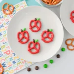 Apple Pretzels that are bow pretzels covered in red melting chocolate with a brown and green M&M for the stem and leaf