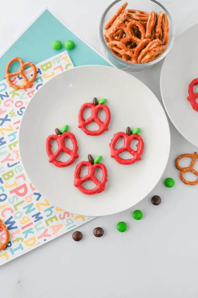 Apple Pretzels that are bow pretzels covered in red melting chocolate with a brown and green M&M for the stem and leaf