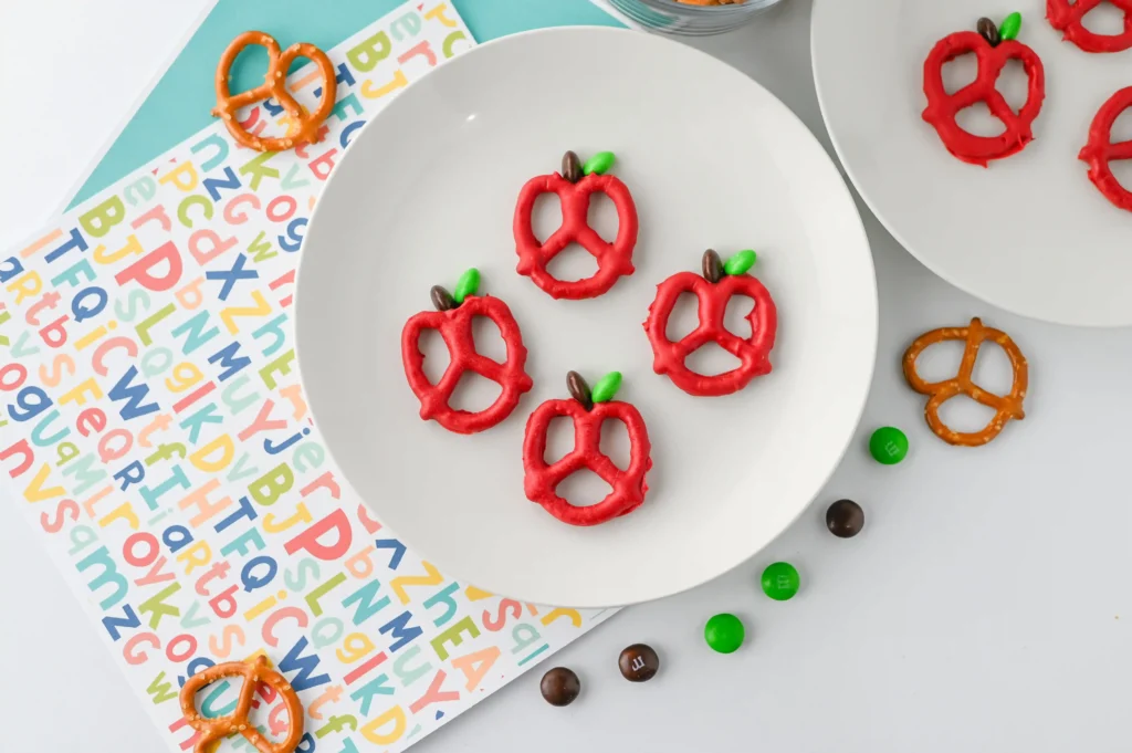 Apple Pretzels that are bow pretzels covered in red melting chocolate with a brown and green M&M for the stem and leaf