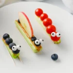 Playful fruit-topped pastry skewers on a white plate; green bases topped with blueberries, an apple slice, and candy eyes for a fun look.