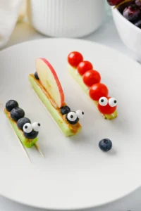 Playful fruit-topped pastry skewers on a white plate; green bases topped with blueberries, an apple slice, and candy eyes for a fun look.