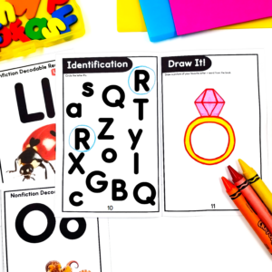 Close-up of children's alphabet learning cards with bold black letters and headings 'Identification' and 'Draw It!', surrounded by colorful crayons and a pink gem illustration on the right.