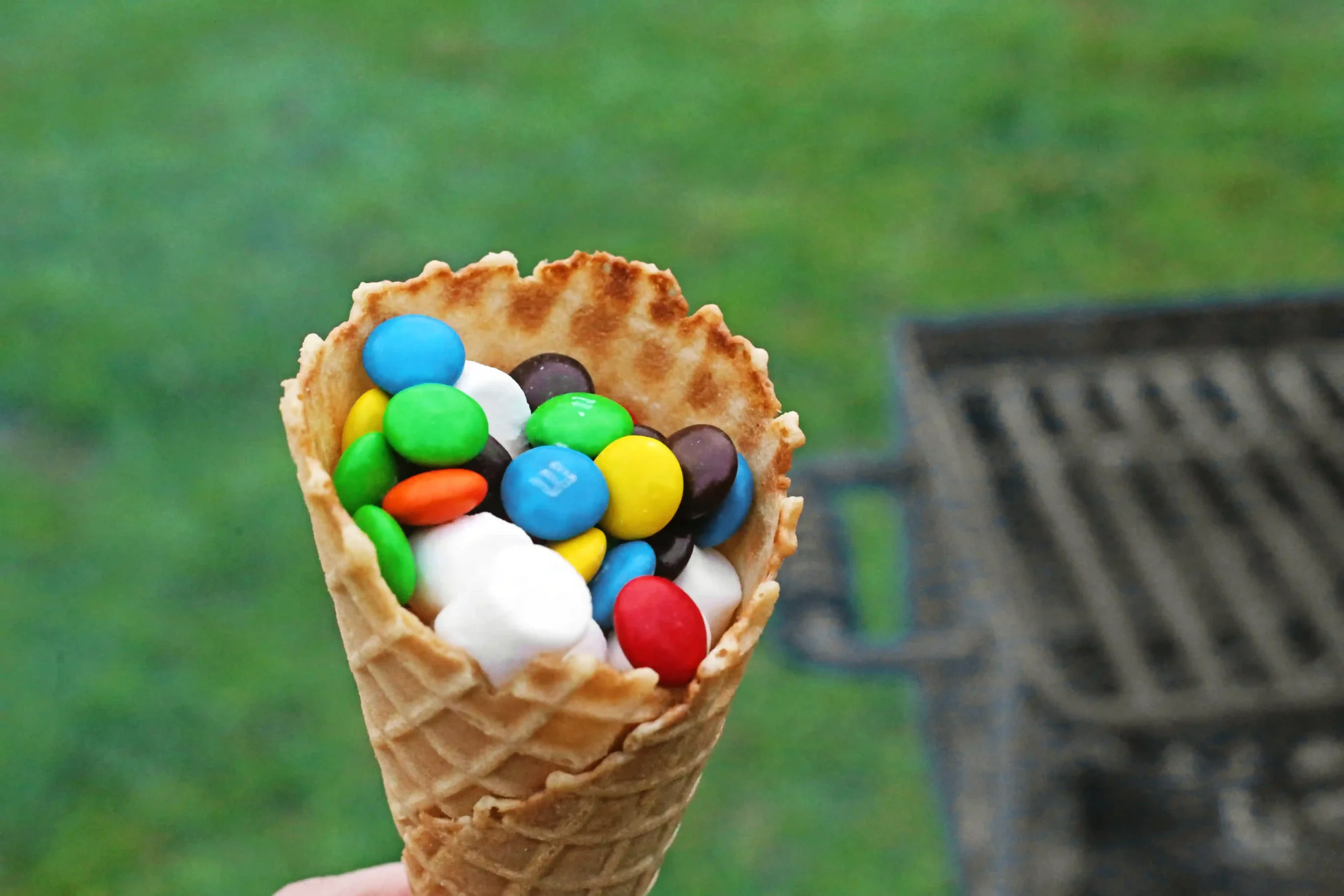 Ice cream cone filled with colorful candy-coated chocolates and whipped cream, held up outdoors against a green background.