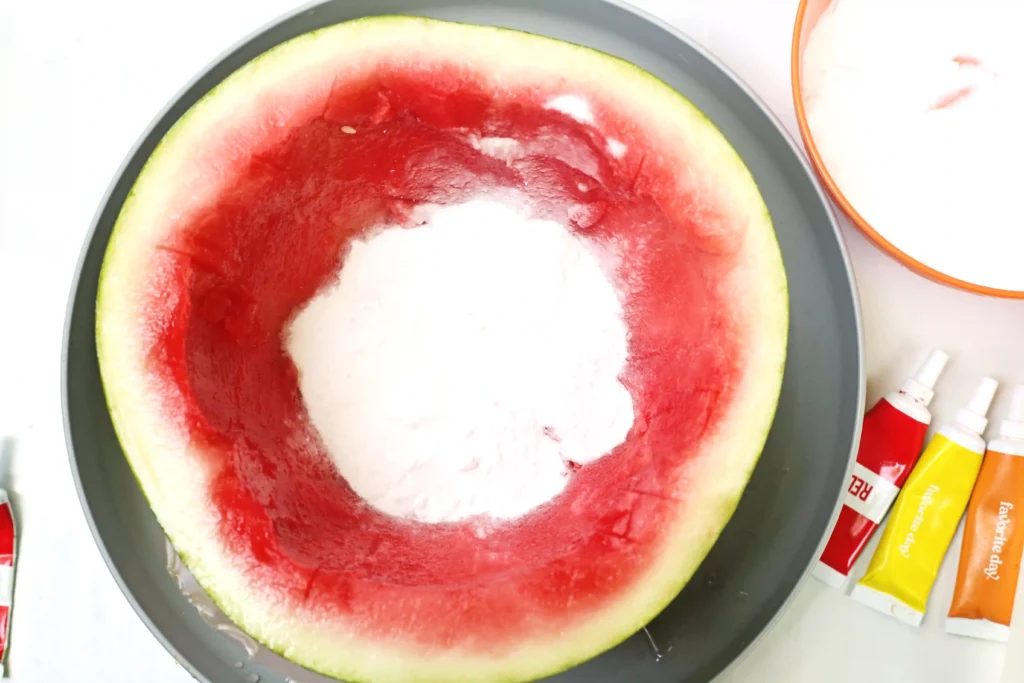 Top-down view of a hollow watermelon ring resting in a dark round dish, with baking soda in the center and food coloring to the right.